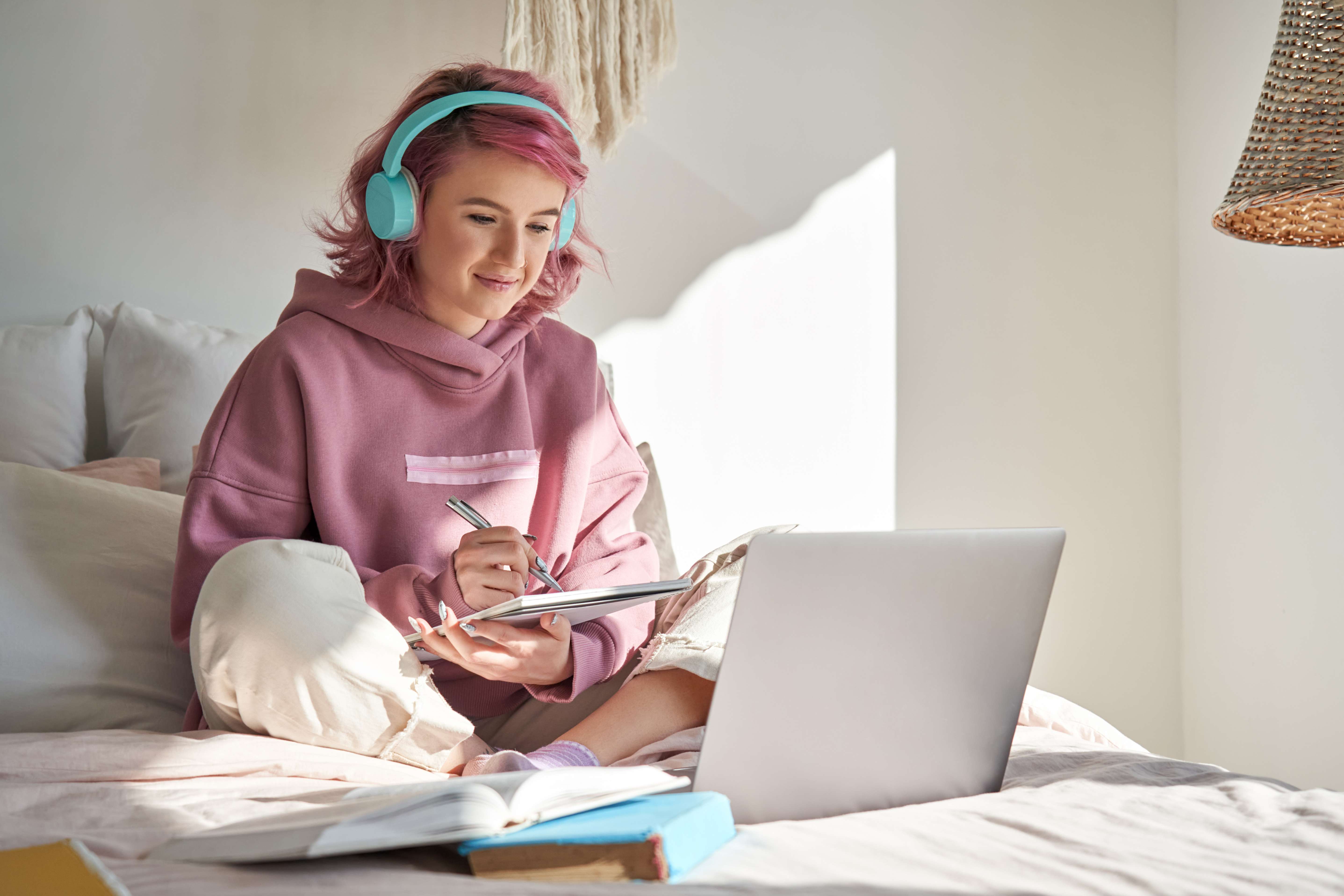 Teen girl with pink hair sitting on a bed, wearing headphones, writing notes and watching laptop.