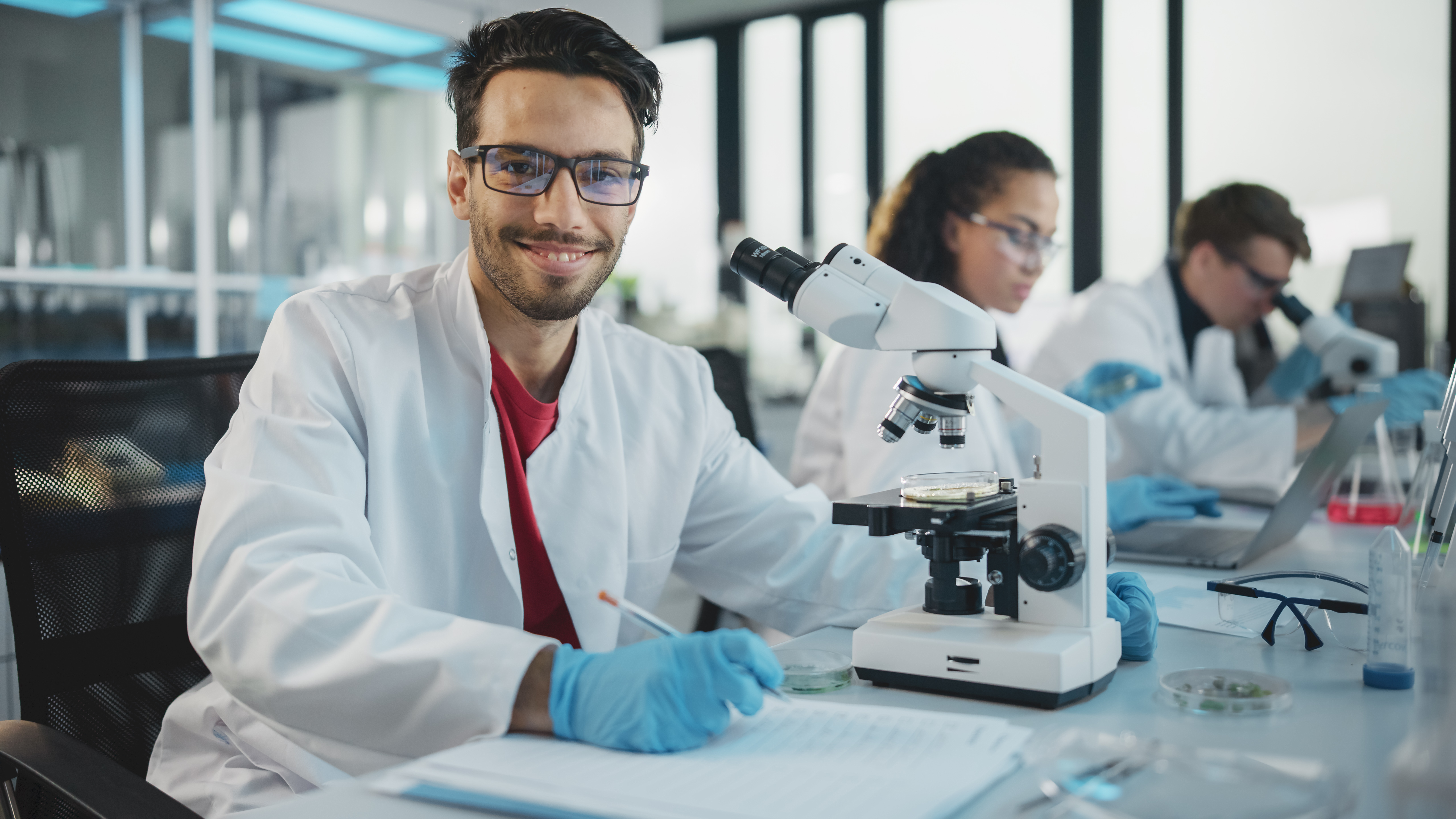 Scientist in lab analyzing samples with microscope, looking at camera and smiling