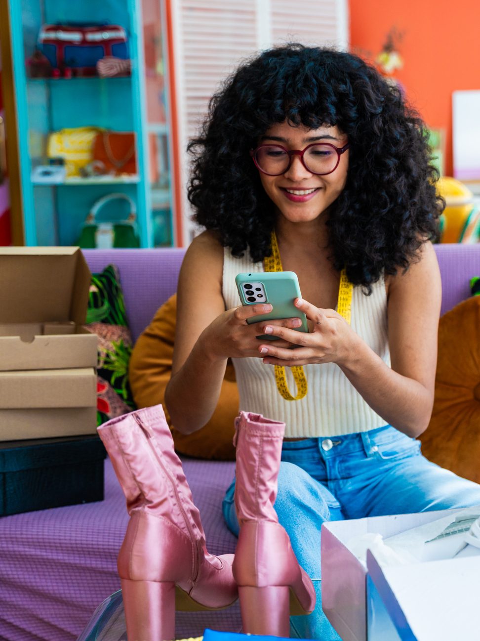 Young adult hispanic woman using a smartphone for her e-commerce clothing business from home, selling pre-loved fashion items and preparing orders for shipping